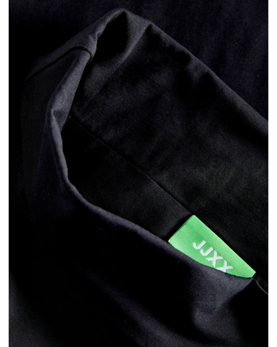 Sort stof med grøn etiket viser mærketeksten "JAXX" i hvid. Etiketten er delvist synlig, omgivet af mørkt, foldet tekstil.