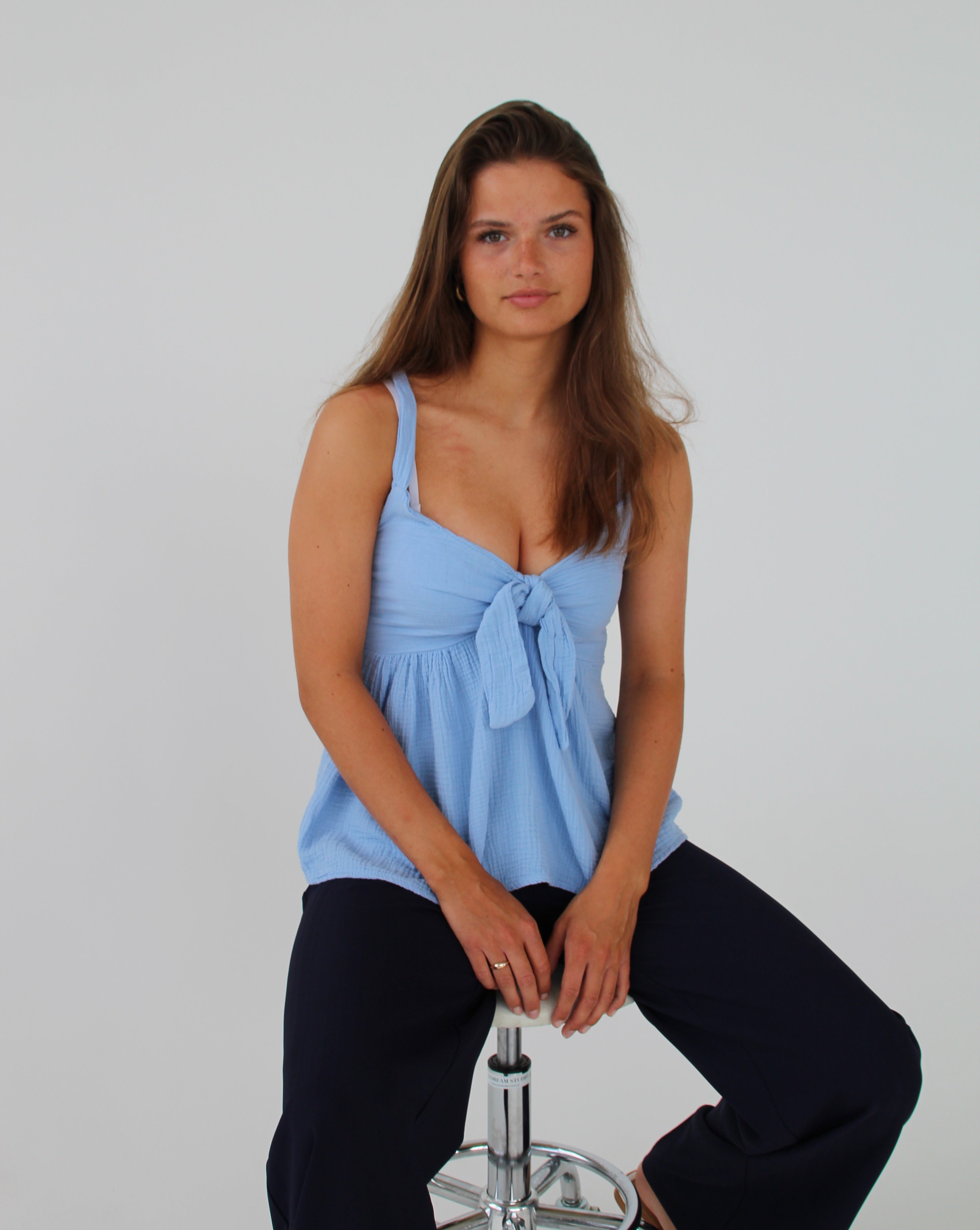 En person sidder på en barstol og kigger fremad. Personen bærer en blå top og mørke bukser i et enkelt studie med en hvid baggrund.