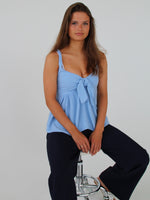 En kvinde sidder afslappet på en barstol. Hun bærer en lyseblå top og mørke bukser i et neutralt rum med hvid baggrund.