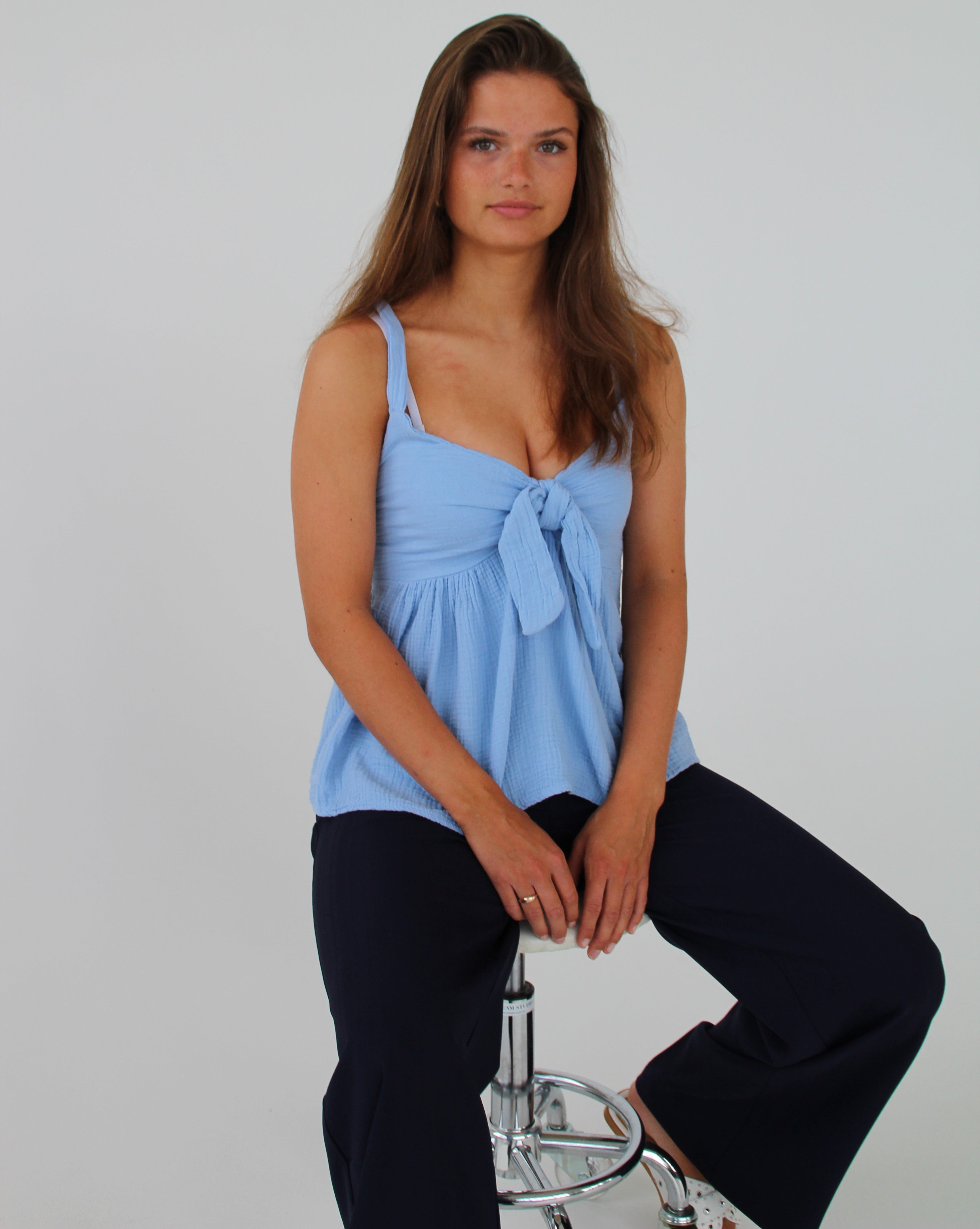 En kvinde sidder afslappet på en barstol. Hun bærer en lyseblå top og mørke bukser i et neutralt rum med hvid baggrund.