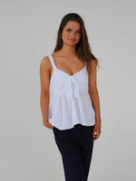 En kvinde iført en hvid top med en sløjfe foran og sorte bukser står foran en neutral hvid baggrund. Hun har hånden i bukselommen.