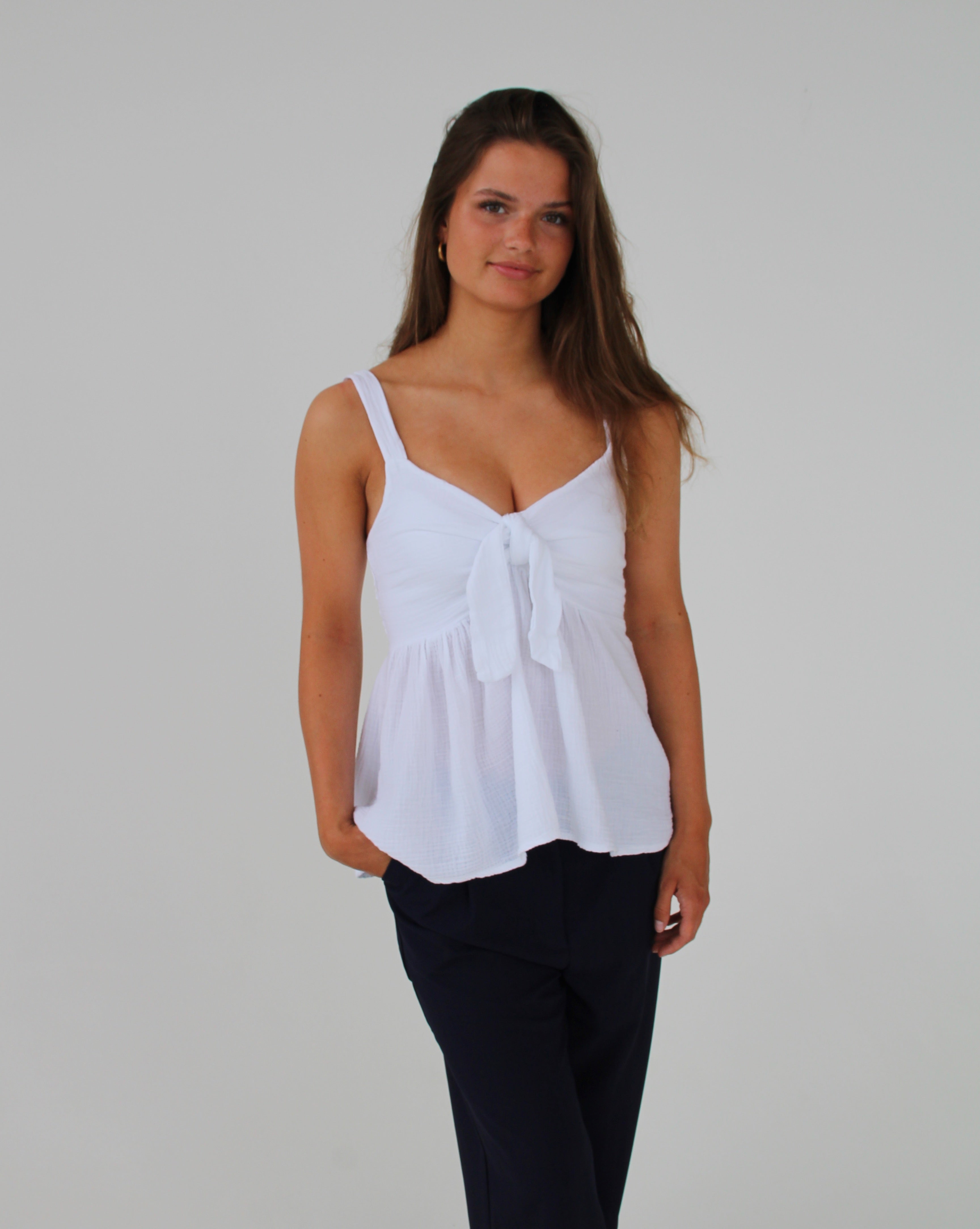 En kvinde iført en hvid top med en sløjfe foran og sorte bukser står foran en neutral hvid baggrund. Hun har hånden i bukselommen.