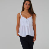 Kvinde iført hvid top står roligt, med hænderne langs siden, på en neutral, hvid baggrund. Hun har løst, langt hår og sorte bukser.