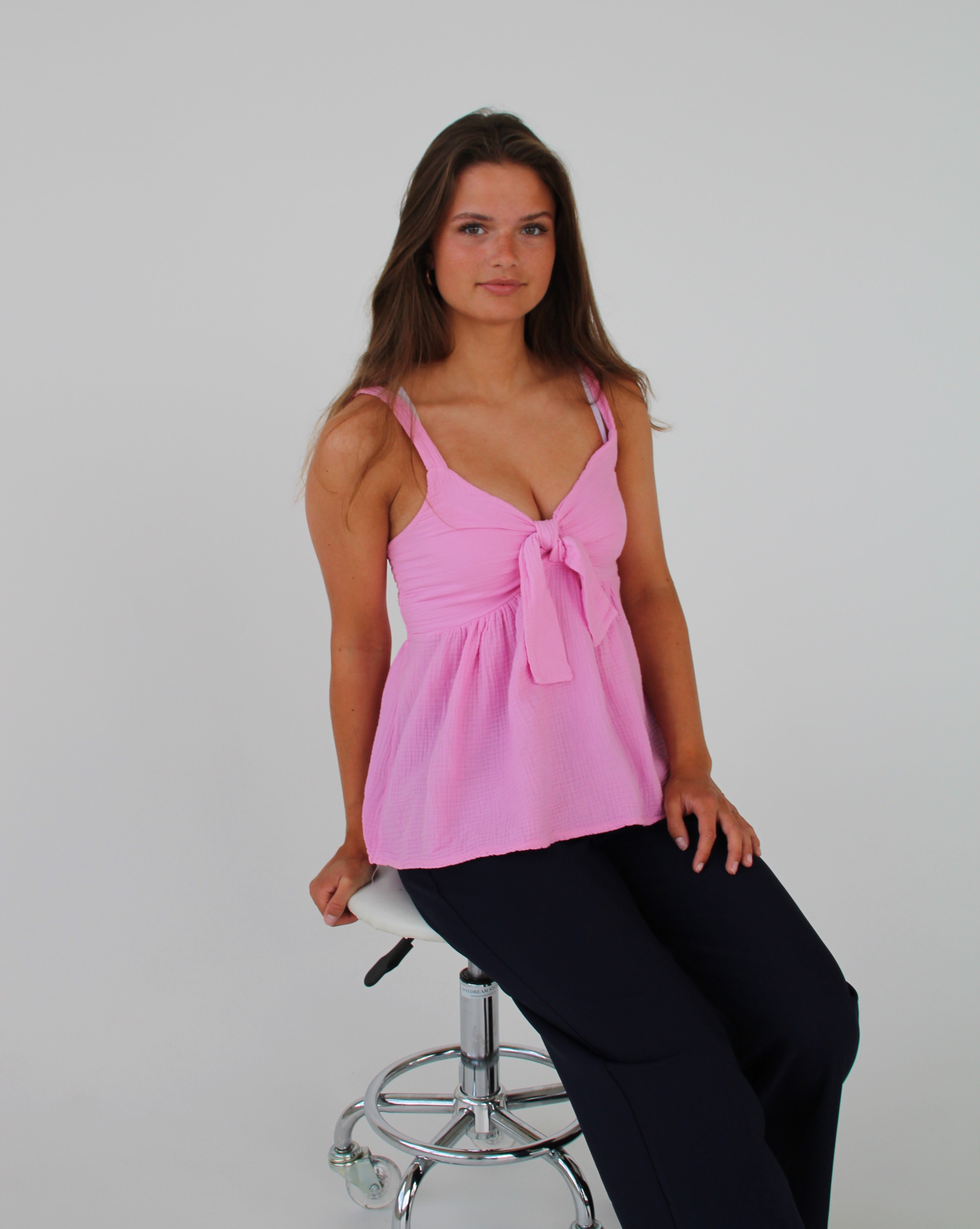 En person sidder stille på en hvid, metal-stol, iført en lyserød top og sorte bukser, med hvid baggrund.