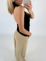 En person tager et spejlbillede iført en sort stropløs top og lysebrune bukser. Personen står sidelæns med lyse baggrund og holder en telefon.