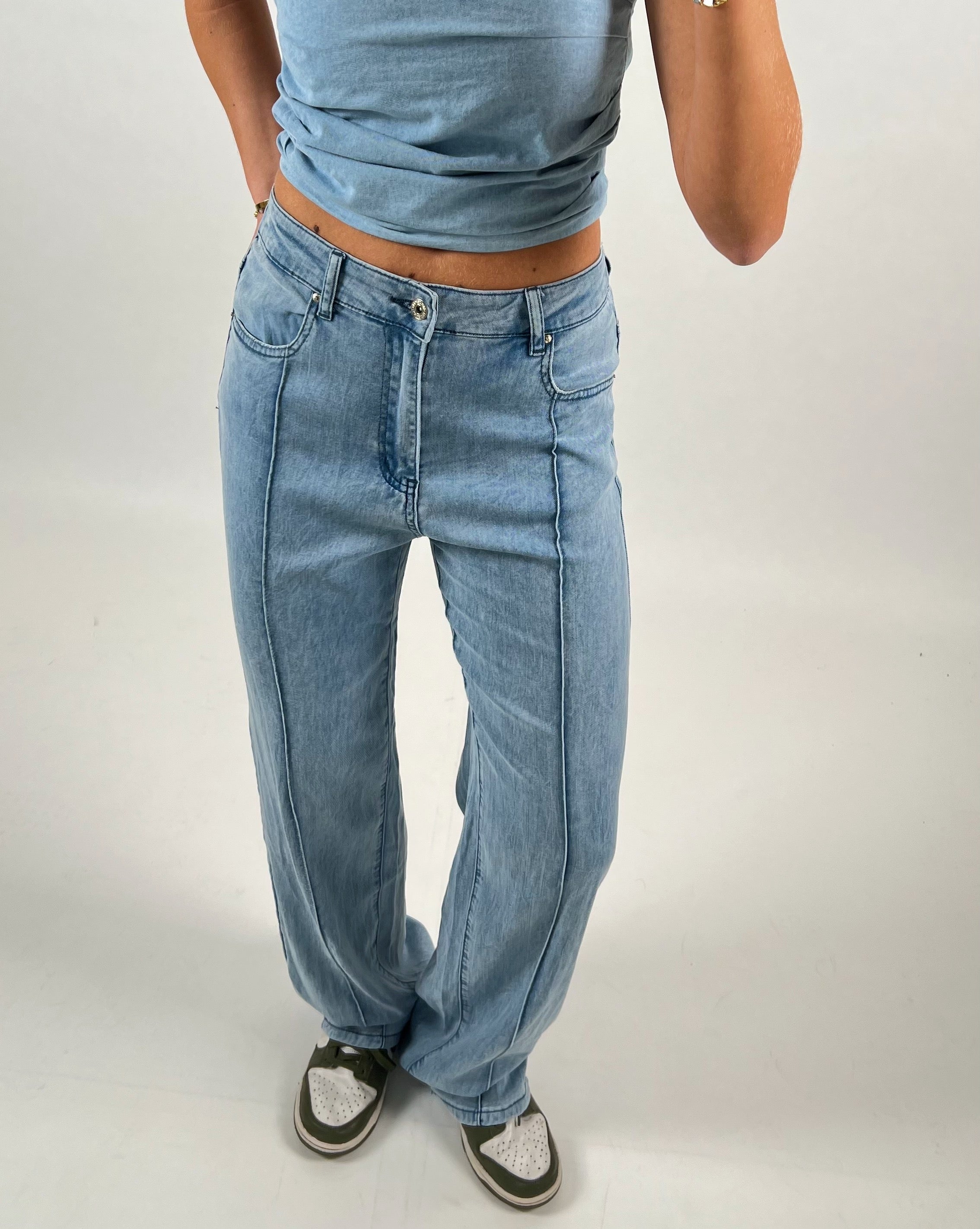 Person iført lyseblå jeans og blå top står med hænderne i lommerne. Personen har grønne og hvide sneakers på en neutral baggrund.