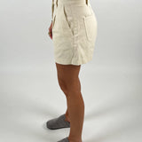 En person iført beige shorts står med siden til, iført grå hjemmesko. Personen har en hvid top på, og der ses en arm med armbånd. Baggrunden er hvid.