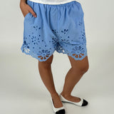 Blå shorts med blomsterudskæringer bæres af en person, der står oprejst. Personen har hænderne i lommerne. En hvid overdel og sko komplementerer det lyse baggrund.