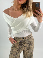 En person med hvid bluse og snakeskin-mønstrede bukser tager et spejlbillede med en smartphone i brun etui. Personen bærer guldsmykker og står i en neutral baggrund.