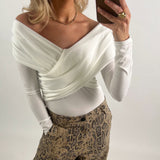 Kvinde i off-shoulder, hvid top tager en selfie med en smartphone. Hun bærer slangeskindsbukser og smykker, står i et neutralt, indendørs miljø.