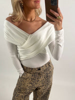 Kvinde i off-shoulder, hvid top tager en selfie med en smartphone. Hun bærer slangeskindsbukser og smykker, står i et neutralt, indendørs miljø.