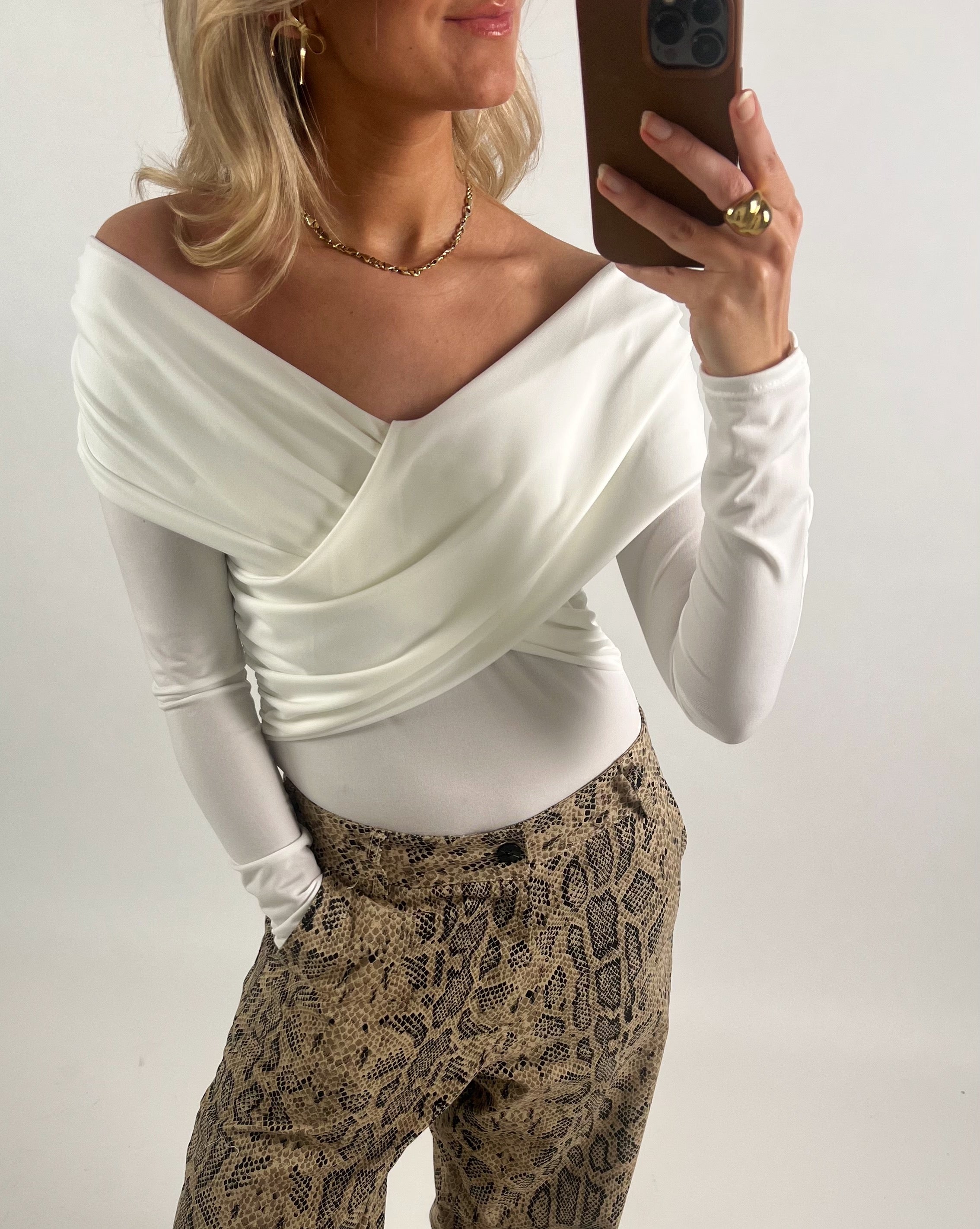 Kvinde i off-shoulder, hvid top tager en selfie med en smartphone. Hun bærer slangeskindsbukser og smykker, står i et neutralt, indendørs miljø.