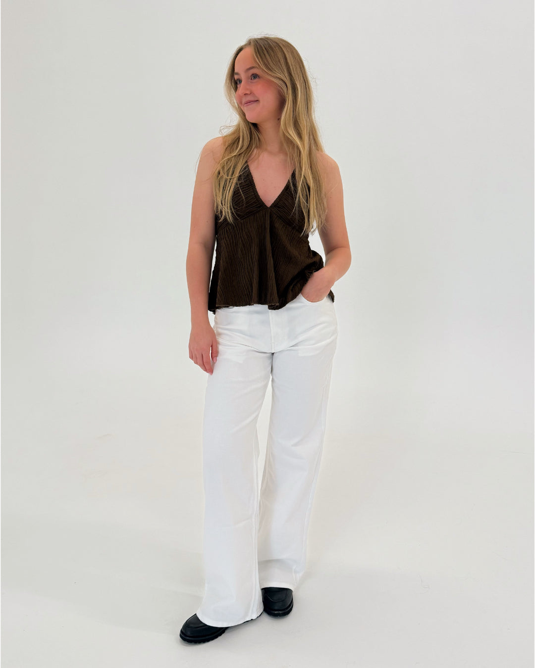 En kvinde står afslappet med hånden i bukselommen. Hun har en brun top, hvide bukser og sorte sko. Baggrunden er enkel og hvid.