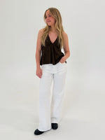 En kvinde står afslappet med hånden i bukselommen. Hun har en brun top, hvide bukser og sorte sko. Baggrunden er enkel og hvid.