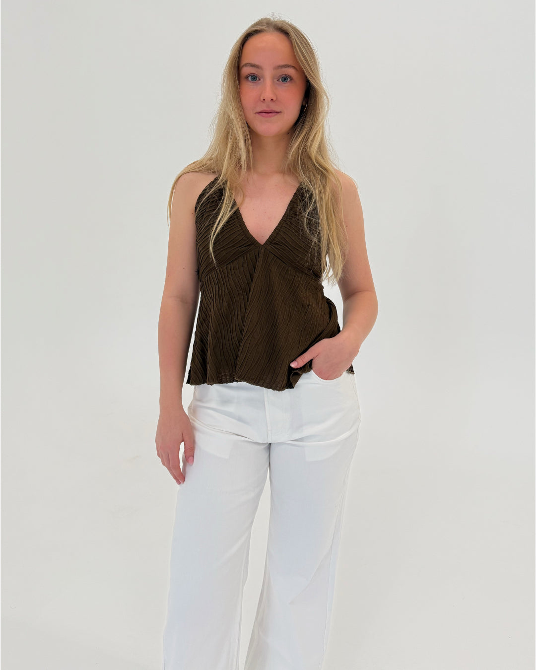 En person står stille iført en brun top og hvide bukser. Personen hviler en hånd i bukselommen i et lyst, minimalistisk rum.