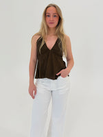 En person står stille iført en brun top og hvide bukser. Personen hviler en hånd i bukselommen i et lyst, minimalistisk rum.