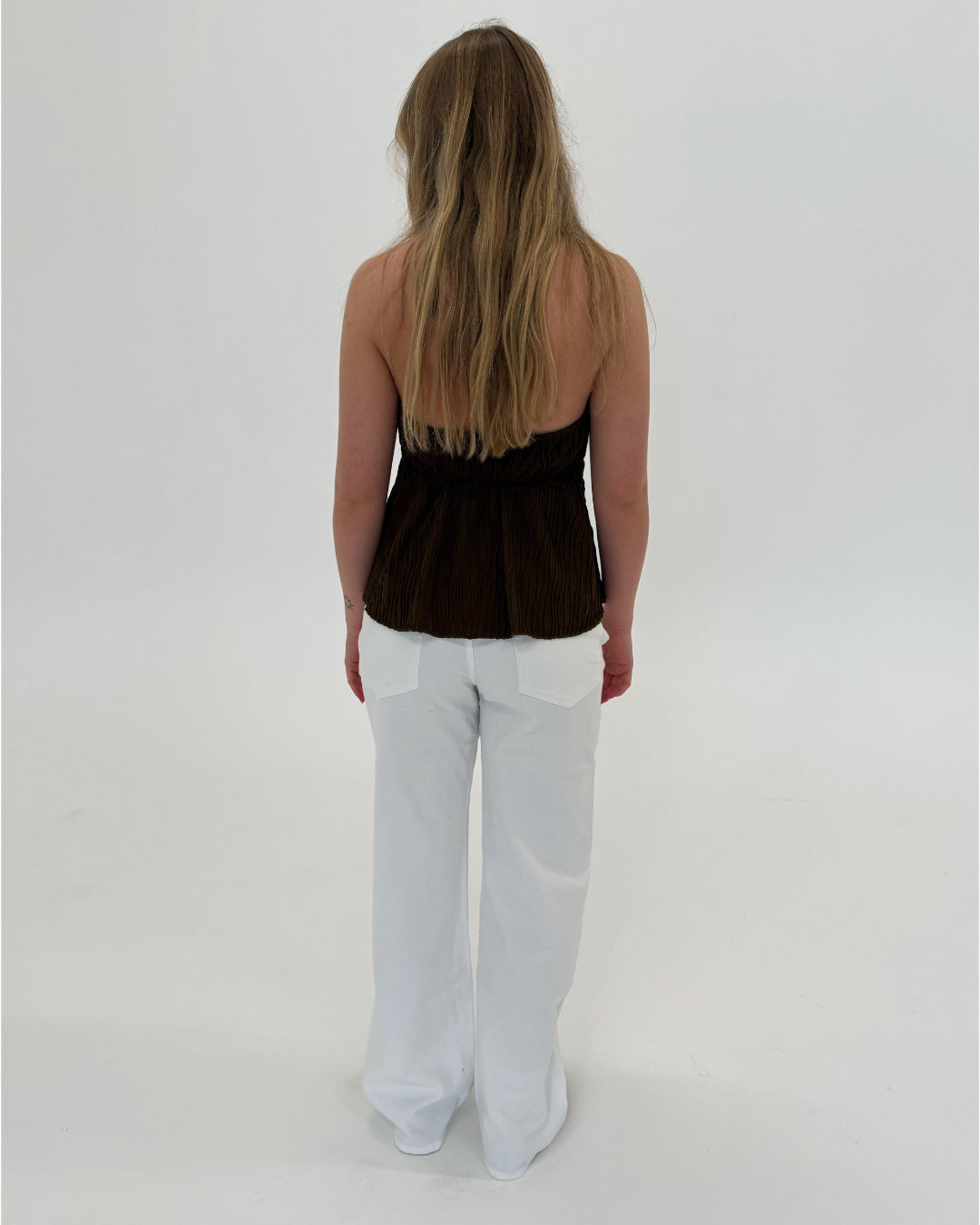 En person står med ryggen til, iført en stropløs brun top og hvide bukser, i et lyst studie uden synlige møbler eller dekorationer.