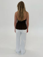 En person står med ryggen til, iført en stropløs brun top og hvide bukser, i et lyst studie uden synlige møbler eller dekorationer.