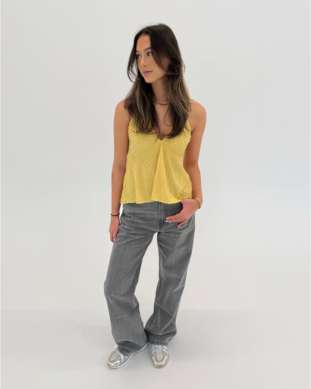 En kvinde står afslappet iført en gul top og grå bukser, med hånden i lommen. Hun befinder sig i et lyst, minimalistisk rum.
