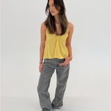 En kvinde står afslappet iført en gul top og grå bukser, med hånden i lommen. Hun befinder sig i et lyst, minimalistisk rum.