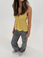 En person står iført en gul top og grå bukser, med hænderne afslappet ved siden, i et lyst, neutralt rum.