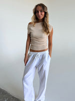 En person står afslappet iført en beige off-shoulder top og hvide bukser. Personen har hænderne i lommerne og befinder sig i et lyst, minimalistisk rum.