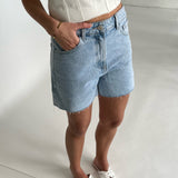 En person iført lyseblå denimshorts med en hånd i lommen står afslappet i et lyst interiør. Personen bærer en hvid top og hvide sandaler.