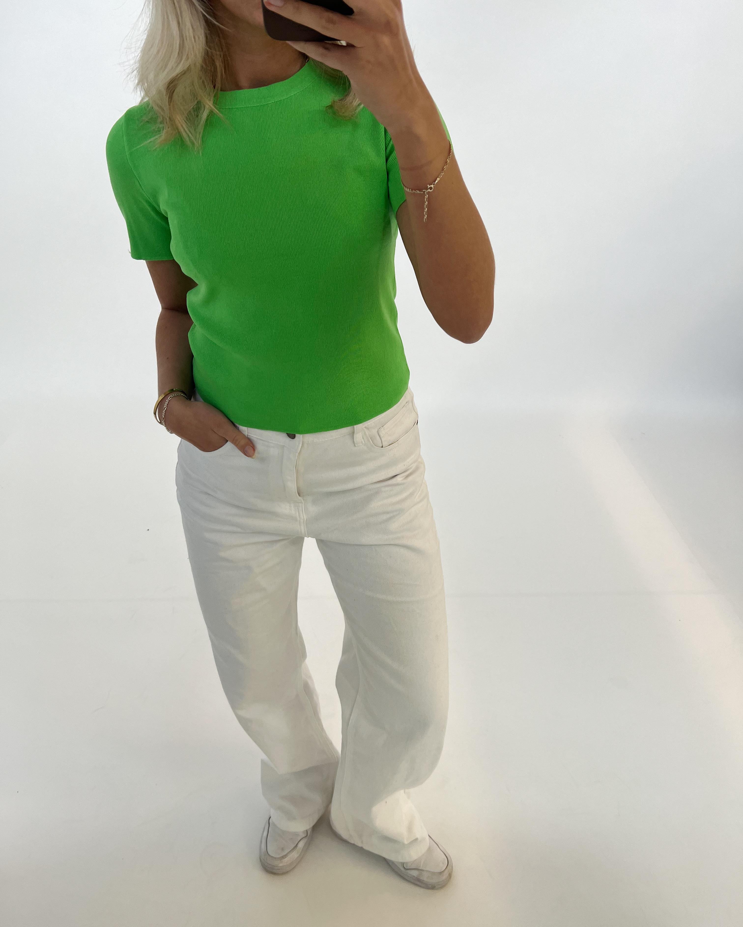 En person iført en grøn t-shirt og hvide bukser tager et spejlbillede i et lyst rum. Personen har en hånd i lommen og en mobiltelefon i den anden hånd.