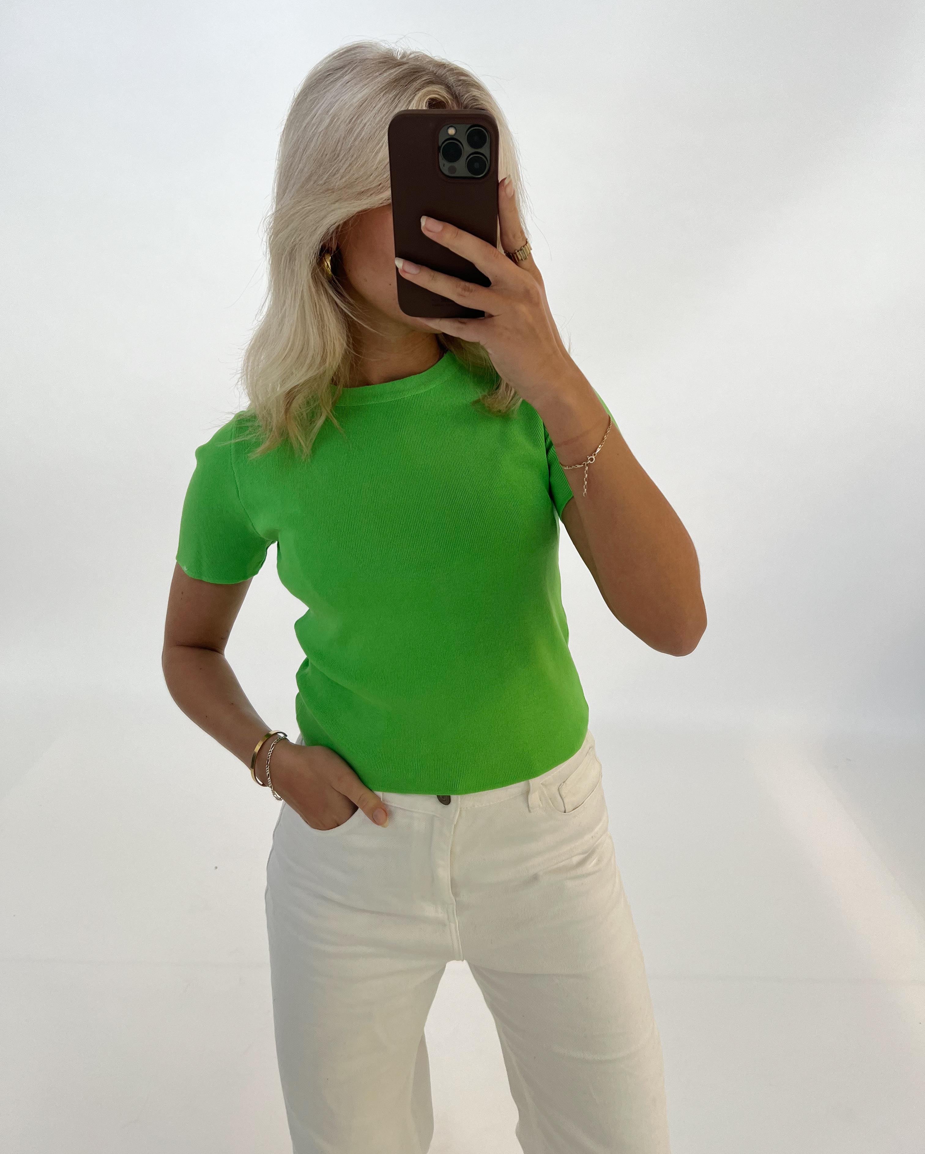 Person tager et spejlbillede, iført grøn T-shirt og hvide bukser. Personen dækker ansigtet med en mobiltelefon i en minimalistisk, lys baggrund.