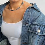 En person bærer en denimjakke over en hvid tanktop og en halskæde med blå perler; personen har lys hud og står mod en lys baggrund.