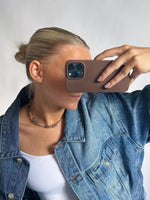En person iført en denimjakke tager et billede med en mobiltelefon. Personen har opsat hår og bærer en hvid top og et perlekæde, set mod en lys baggrund.