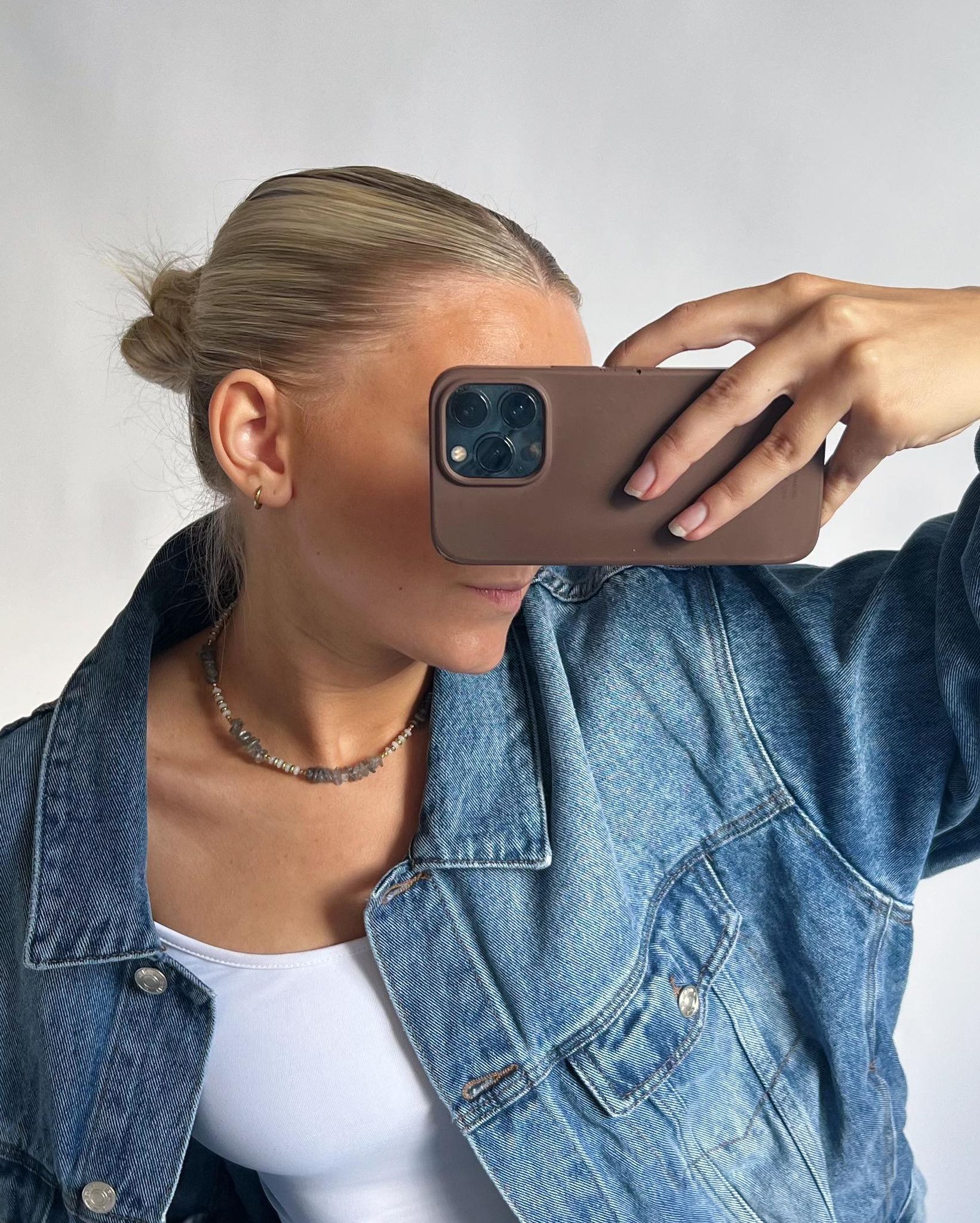 En person iført en denimjakke tager et billede med en mobiltelefon. Personen har opsat hår og bærer en hvid top og et perlekæde, set mod en lys baggrund.