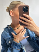 En person holder en telefon i en brun etui foran ansigtet, iført en denimjakke, grøn halskæde, og flere armbånd, i et neutralt rum.