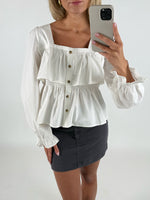 Kvinde iført hvid bluse med knapper, holder en smartphone til at tage et spejlbillede. Baggrunden er neutral. Hun har en grå nederdel på og står i et lyst rum.