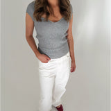 Kvinde iført grå top og hvide bukser står i et lyst rum, med én hånd i bukselommen. Hun har langt, bølget hår og bærer røde sko.