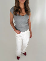 Kvinde iført grå top og hvide bukser står i et lyst rum, med én hånd i bukselommen. Hun har langt, bølget hår og bærer røde sko.