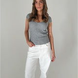 En kvinde står afslappet iført en grå strikket top og hvide bukser, med en neutral baggrund. Hendes ene hånd er i lommen.