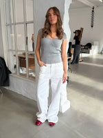 En kvinde iført grå top og hvide bukser står ved en væg i et fotostudie, mens en anden person fotograferer i baggrunden.