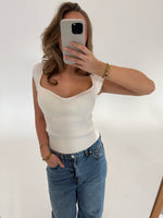 En person tager et spejlbillede med en smartphone, iført en hvid ærmeløs top og blå jeans, i et lyst rum.