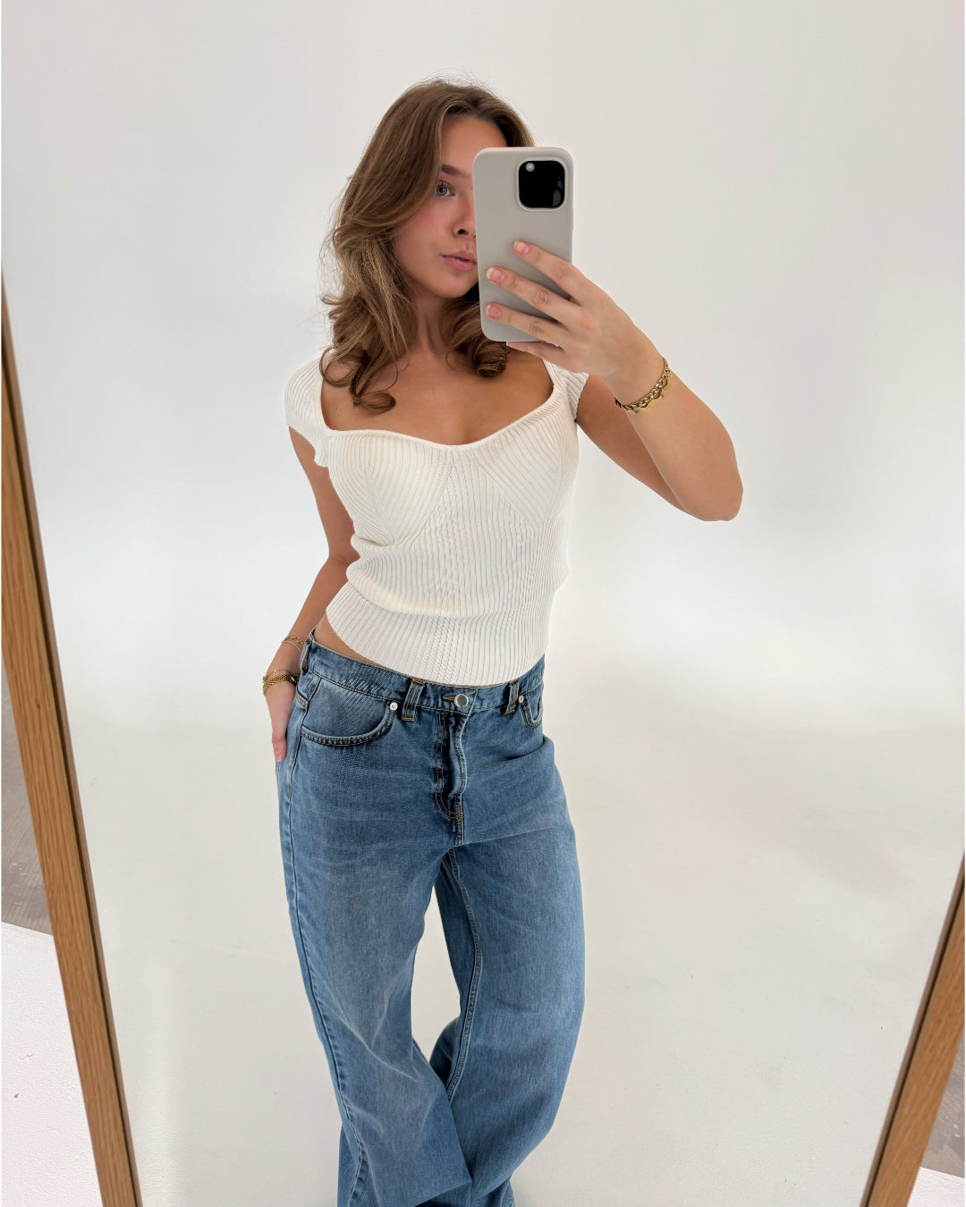 Kvinde tager et spejlbillede med en smartphone iført en hvid top og blå jeans. Hun står i et minimalistisk rum med hvid baggrund, træramme synlig på spejlets kant.