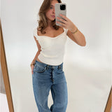 Kvinde tager et spejlbillede med en smartphone iført en hvid top og blå jeans. Hun står i et minimalistisk rum med hvid baggrund, træramme synlig på spejlets kant.