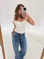 Kvinde tager et spejlbillede med en smartphone iført en hvid top og blå jeans. Hun står i et minimalistisk rum med hvid baggrund, træramme synlig på spejlets kant.