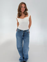 Kvinde står afslappet i hvid top og blå jeans i et lyst rum. Hun har hånden i lommen og blikket rettet mod kameraet.