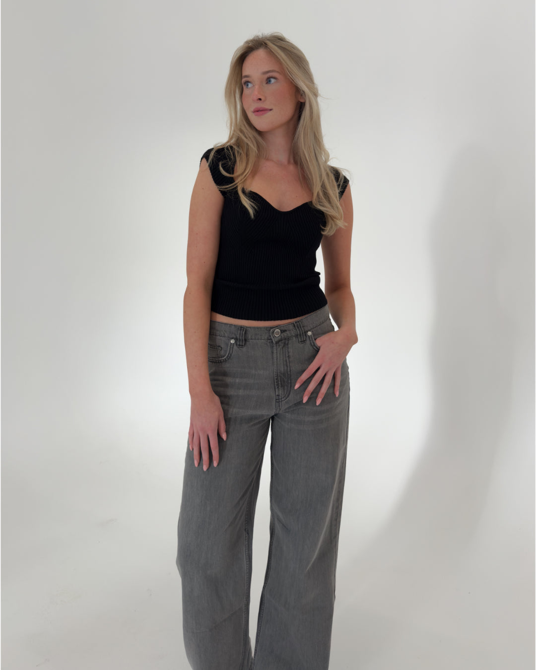 En kvinde står, iført en sort top og grå bukser, ser til siden i et minimalistisk, hvidt rum.