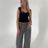 En kvinde står, iført en sort top og grå bukser, ser til siden i et minimalistisk, hvidt rum.