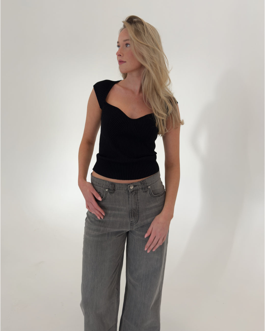 En kvinde iført sort top og grå jeans står afslappet med hænderne i lommerne i et hvidt studie.