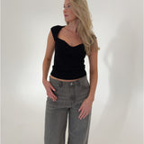 En kvinde iført sort top og grå jeans står afslappet med hænderne i lommerne i et hvidt studie.