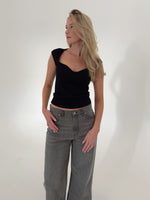 En kvinde iført sort top og grå jeans står afslappet med hænderne i lommerne i et hvidt studie.