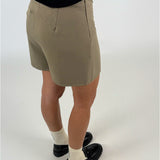 En person iført beige shorts står oprejst, iført sorte sko og hvide strømper, på en lys baggrund. Personen bærer også en sort trøje.