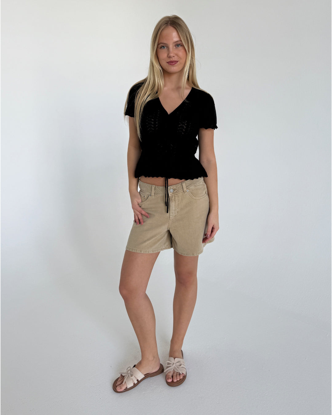 Kvinde står afslappet, iført sort bluse og beige shorts med sløjfesandaler, i et rum med hvid baggrund. Hun har langt lyst hår og smiler let.